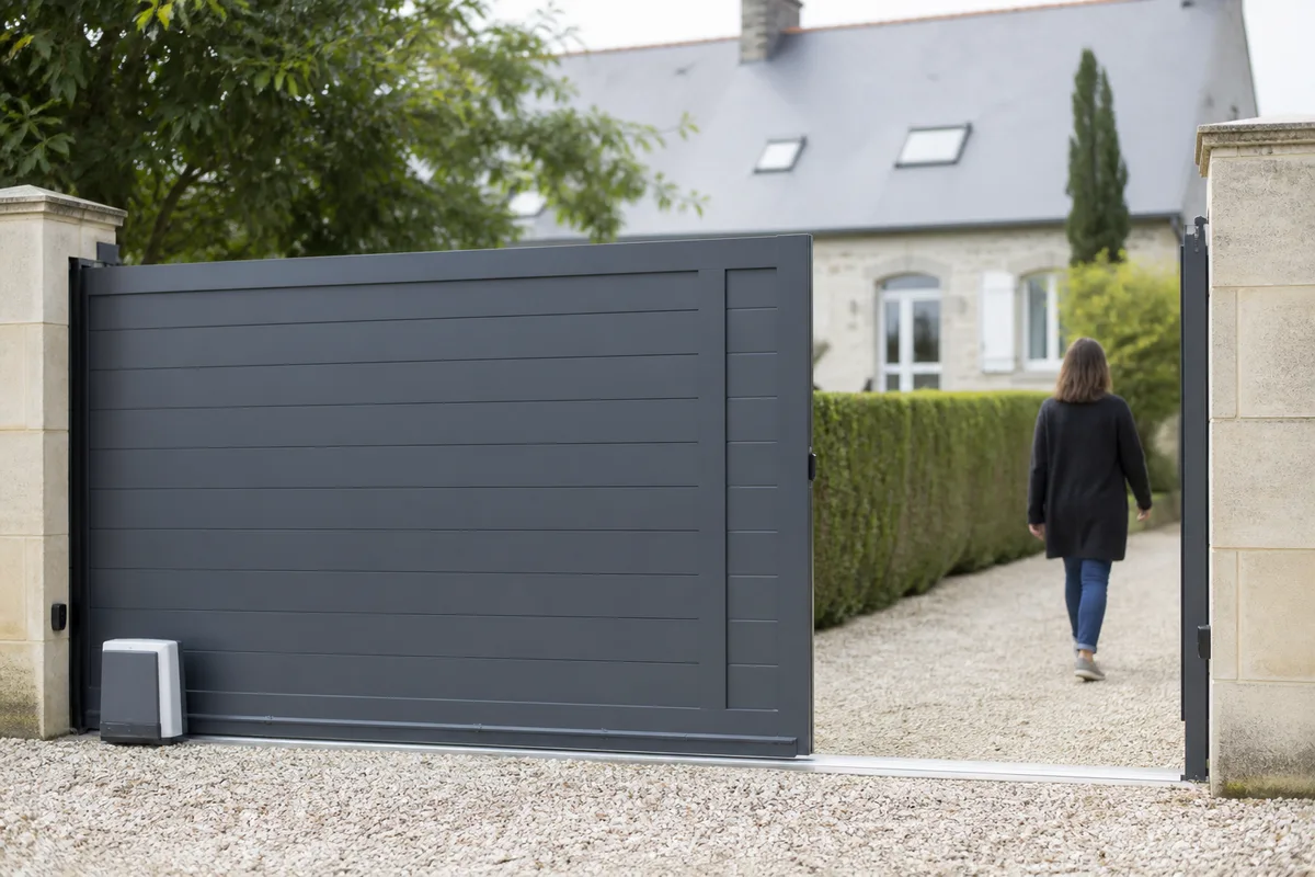 Portail aluminium coulissant gris anthracite devant un pavillon, allée en gravier et haie taillée